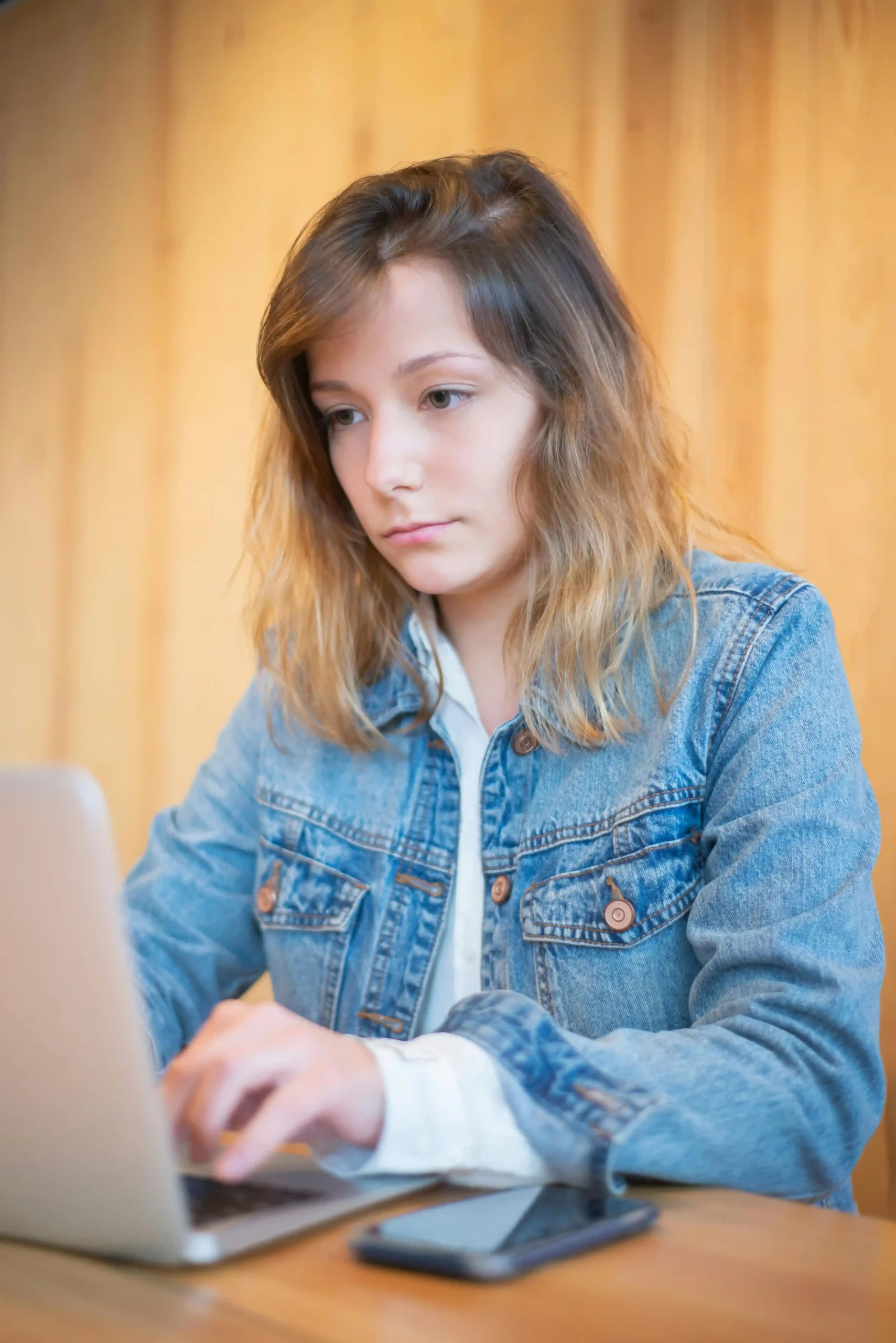 A young woman focused on her laptop, wearing a denim jacket.