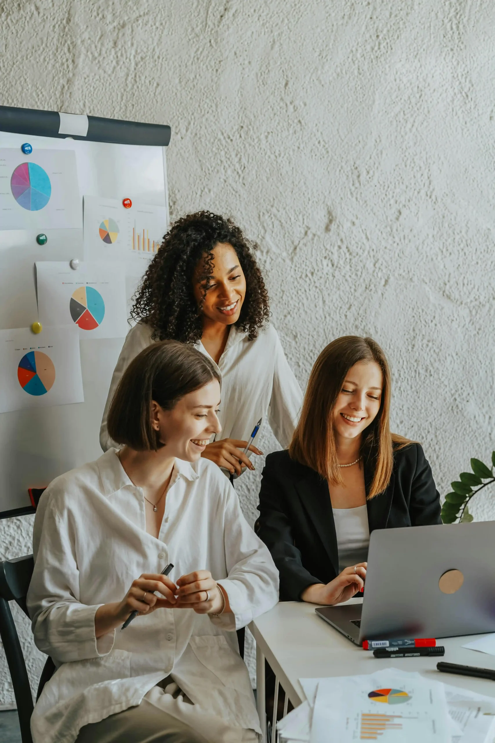 Three women collaborating and smiling while working on a laptop near charts on a board.