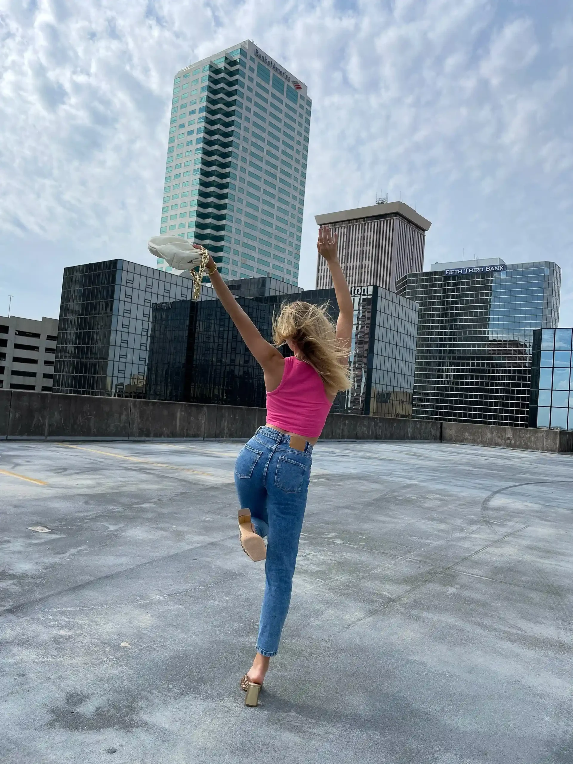 Woman in a pink top and jeans joyfully leaping on a rooftop with city skyscrapers in the background.